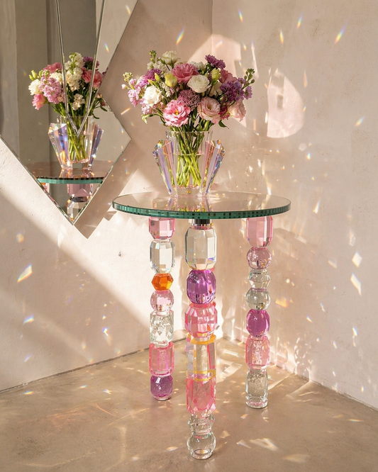 Glass table with colorful cylindrical legs holding flower vases in a room with sunlight filtering through.