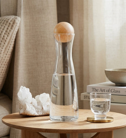 Clear glass carafe with wooden lid on a wooden table, next to a glass of water and decorative items.