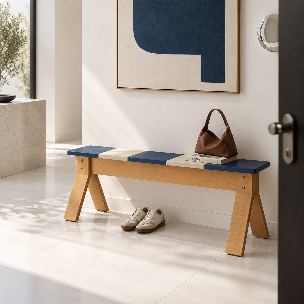 A modern wooden entryway bench with a blue and cream upholstered seat, styled in a bright minimalist hallway with a brown handbag, books, and a pair of beige sneakers underneath.