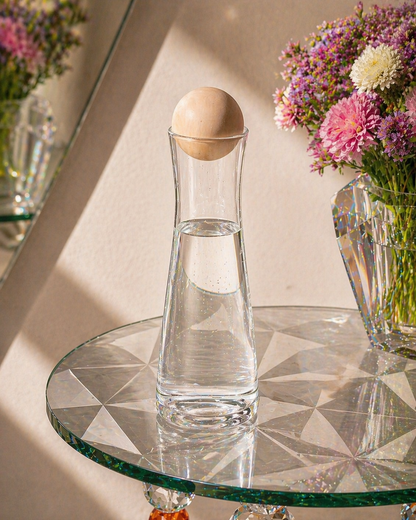 Tapered glass decanter with spherical wooden lid sitting on a refractive geometric glass table with colorful flowers in the background.