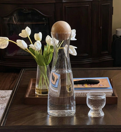 Clear glass carafe with wooden cap on a wooden table with flowers and books in the background