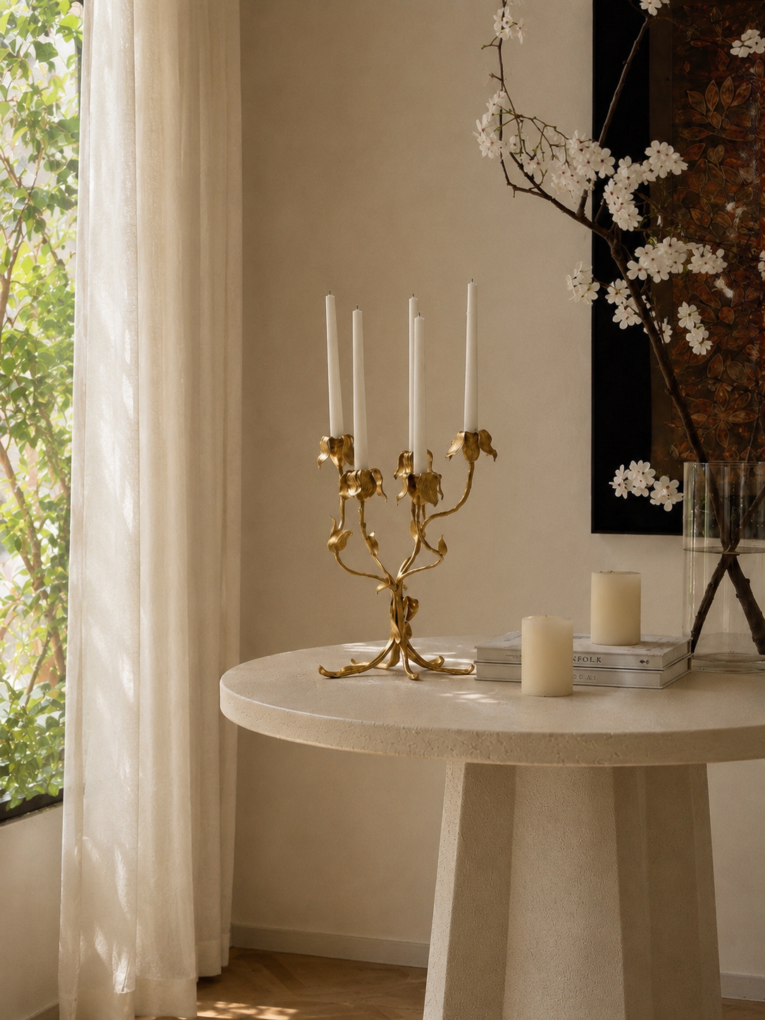 Gold candelabra with candles on a round marble table near a window with sheer curtains.