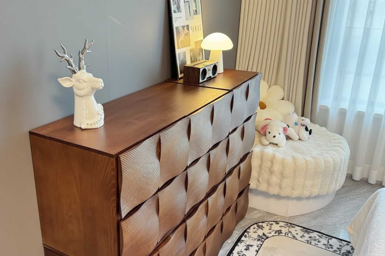 Ausi tall wooden chest of drawers with a wavy, quilted front design, styled in a home setting with a laptop, books, a white deer figurine, and a lamp on top.