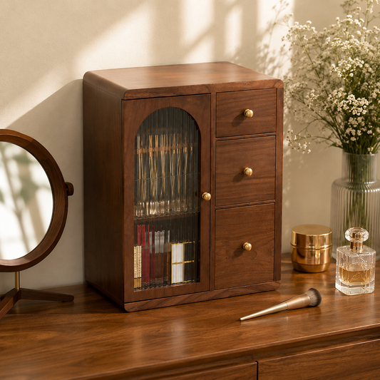 A dark brown wooden vanity organizer cabinet with a glass-front door and three drawers, styled on a wooden table with a mirror, makeup brush, and flowers in the background.