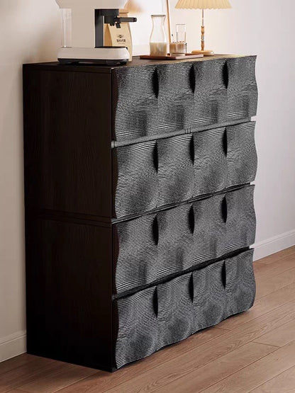 Ausi black wooden chest of drawers with four large front-facing drawers covered in textured gray fabric, styled in a room with a lamp and decor on top, set against a white wall and light wood floor.