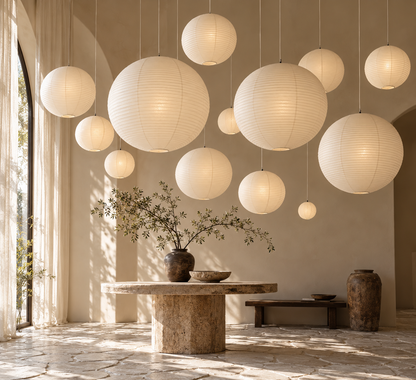 A collection of large, spherical white paper lanterns in various sizes hanging from the ceiling in a sunlit, minimalist modern room with a stone table and vase.