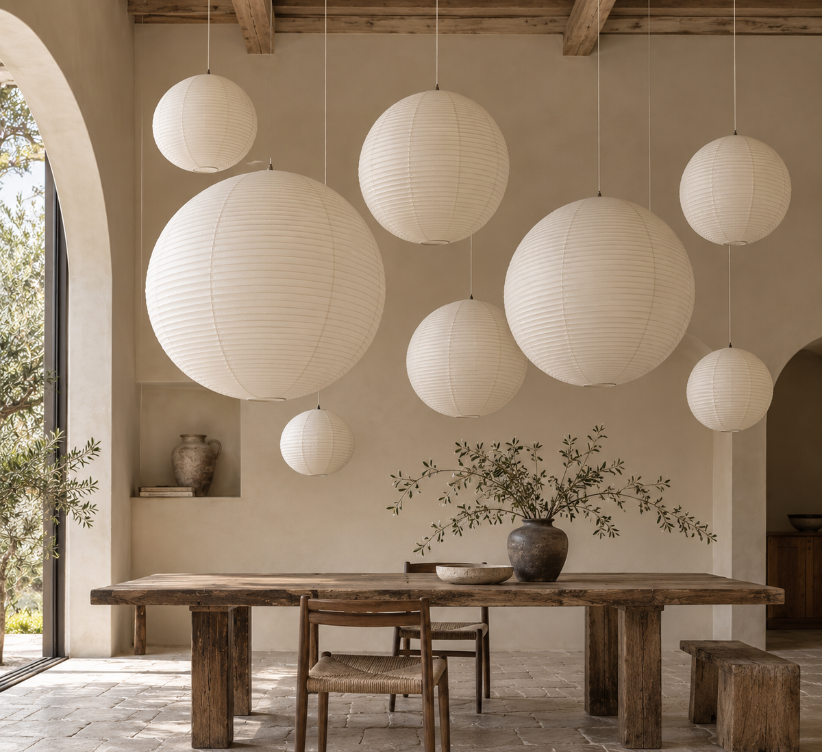 A set of large, off-white paper lantern pendant lights in various sizes hanging from the ceiling of a rustic, minimalist dining room with a wooden table and chairs.