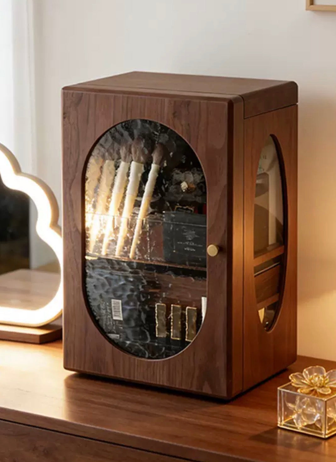 Wooden display cabinet with glass door on a wooden surface, containing makeup brushes and products.